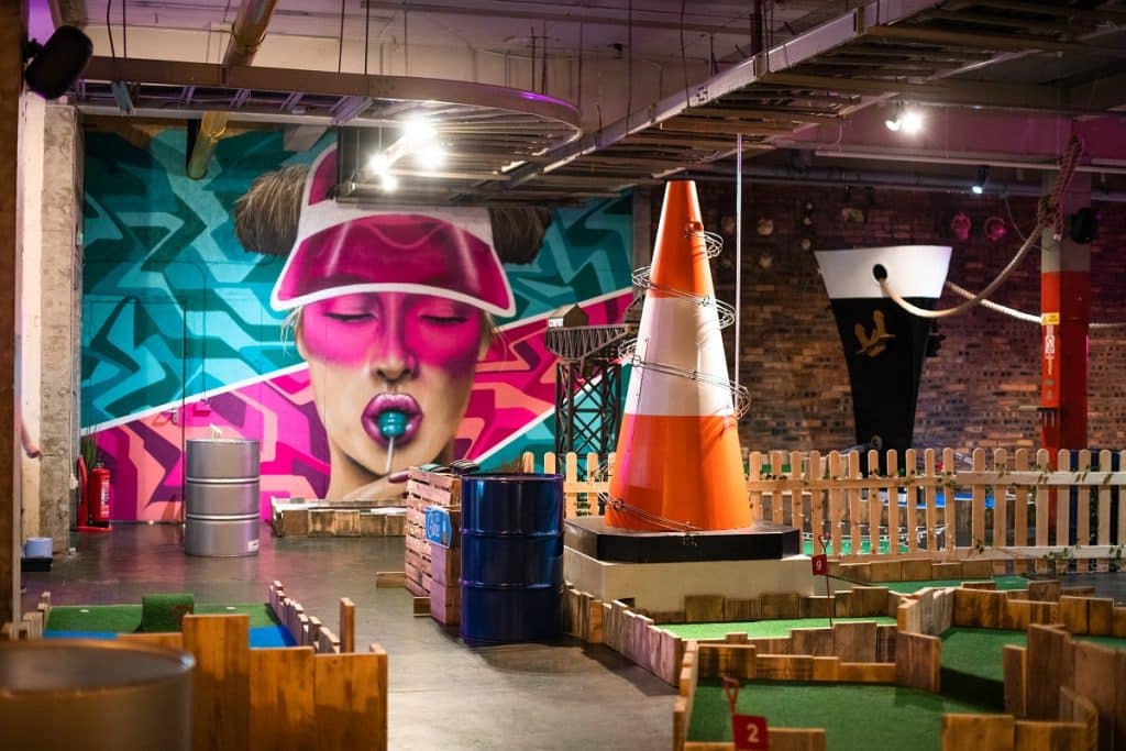 A wide view of Fore Play Crazy Golf, with a giant traffic cone and a mural of a woman eating a lollipop featuring amongst the decor.