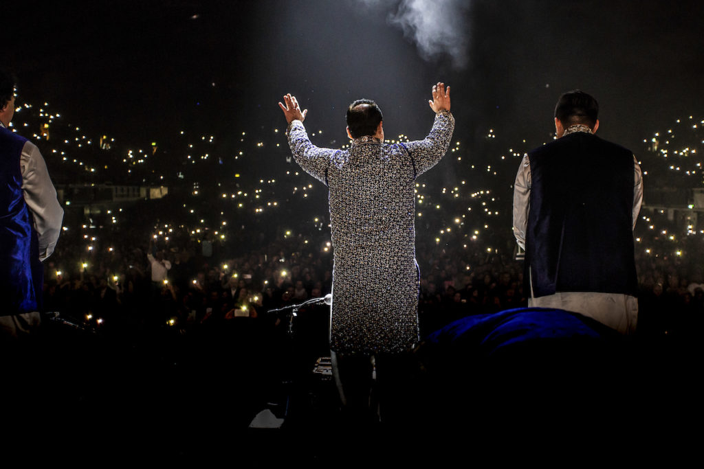 the back of Pakistani singer Rahat Fateh Ali Khan with arms raised in front of a crowd holding lights